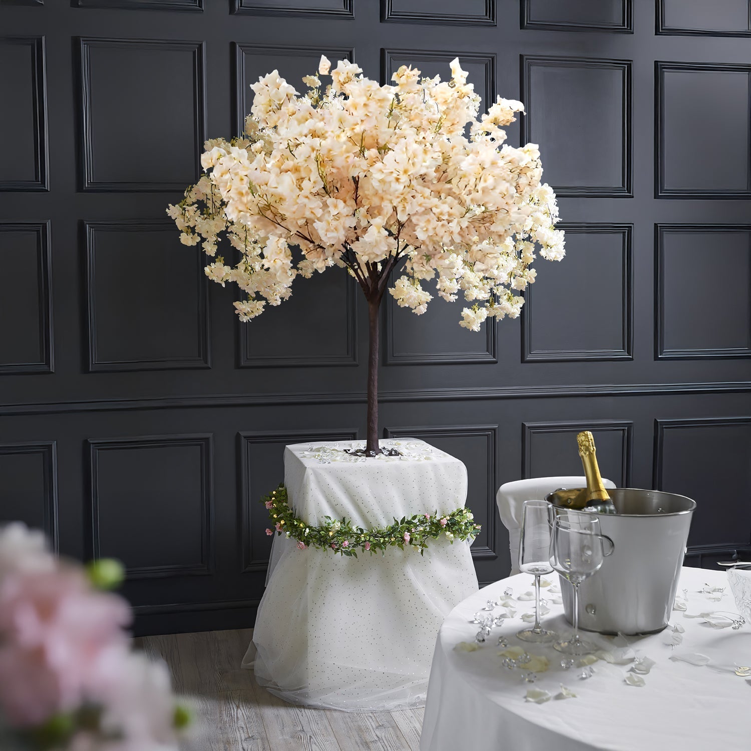 The Prunus Ukon Blossom Tree by Landmark Decor stands on a draped pedestal adorned with a garland. A nearby round table holds two champagne flutes and an ice bucket with champagne, set against dark paneled walls that enhance the elegant scene.