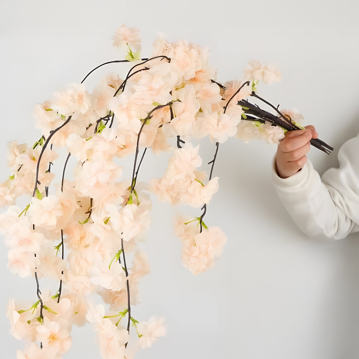 Someone wearing a white long-sleeve shirt holds a bundle of Prunus Ukon Blossom Tree by Landmark Decor, featuring light peach-colored cherry blossoms with beige drooping branches, set against a plain light gray backdrop.