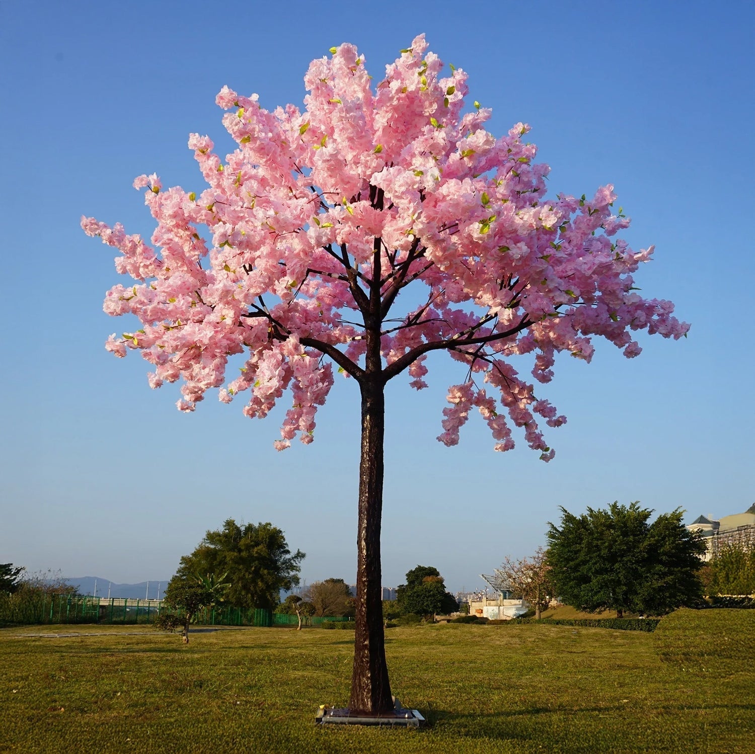 The Romantic Blossom by Landmark Decor depicts a lone cherry blossom tree with rose pink flowers in a lush green field under a clear blue sky, framed by distant trees and a building in the background.