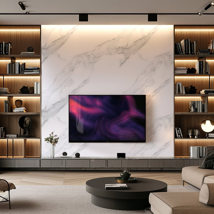 A modern living room features a large wall-mounted TV with abstract art, enhanced by Landmark Decors Pearl Mist Marble Durable PVC Wall panels. Illuminated wooden shelves with books and decor surround it. A round black coffee table and gray sofa complete the cozy design.