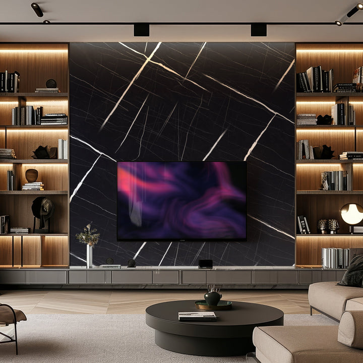 A modern living room features a large TV mounted on Landmark Decors Noir Frost Marble Durable PVC Wall, showcasing white veins for luxury. Built-in wooden shelving with books and decor items flanks the TV. A round black coffee table rests on the light wood floor, surrounded by a gray sofa.