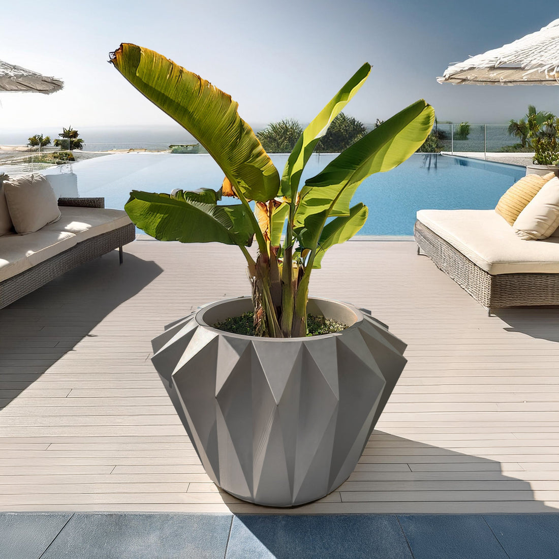 A large, Diamanta Elegant Planter by Landmark Decor, filled with tropical plants, is on a deck near a pool. Wicker lounge chairs and umbrellas sit in the background, while an ocean view enhances the seamless blend of luxury and nature with these exquisite handmade planters.