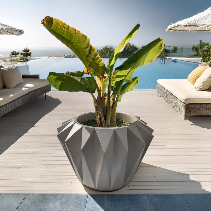 A large, Diamanta Elegant Planter by Landmark Decor, filled with tropical plants, is on a deck near a pool. Wicker lounge chairs and umbrellas sit in the background, while an ocean view enhances the seamless blend of luxury and nature with these exquisite handmade planters.
