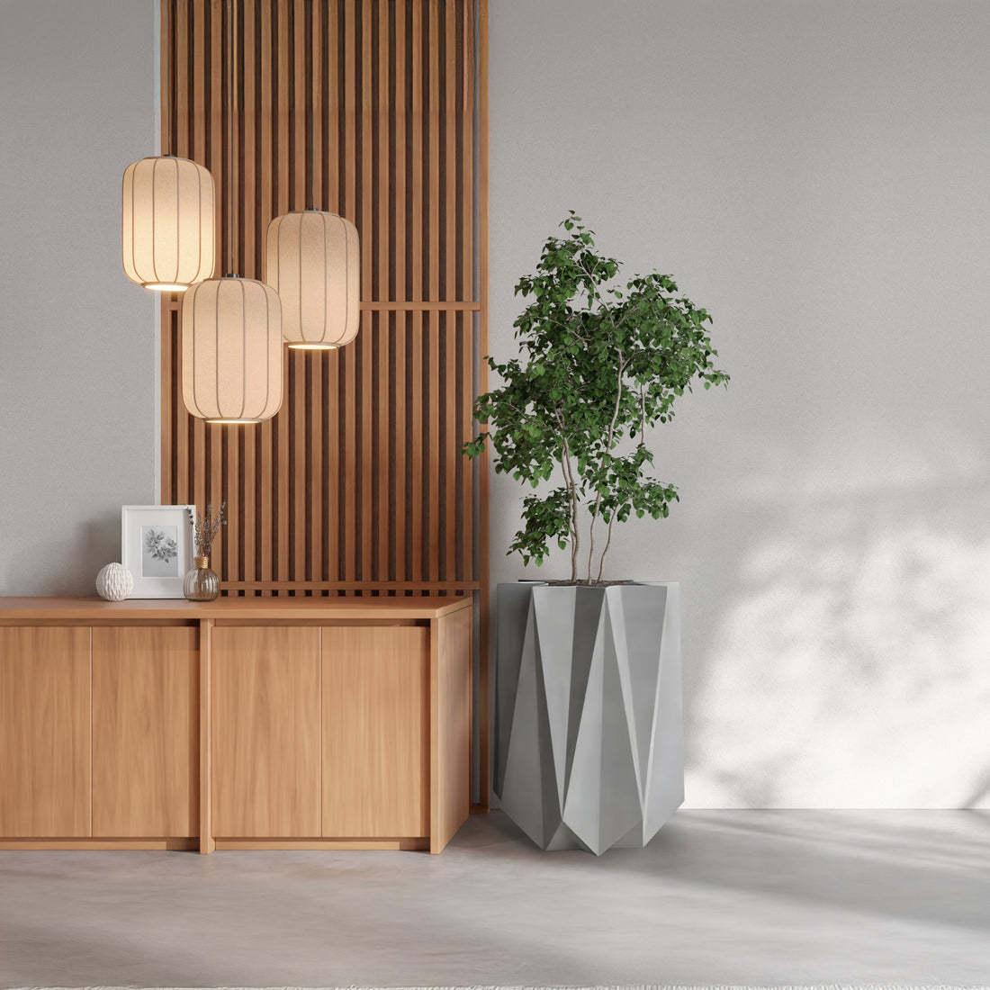 This minimalist interior exudes luxury with wooden paneling and three hanging lanterns. A wooden cabinet displays a framed picture and decor pieces, while the Visione Astratta Elegant Planter by Landmark Decor adds natures touch, casting shadows on the light-colored wall and floor.