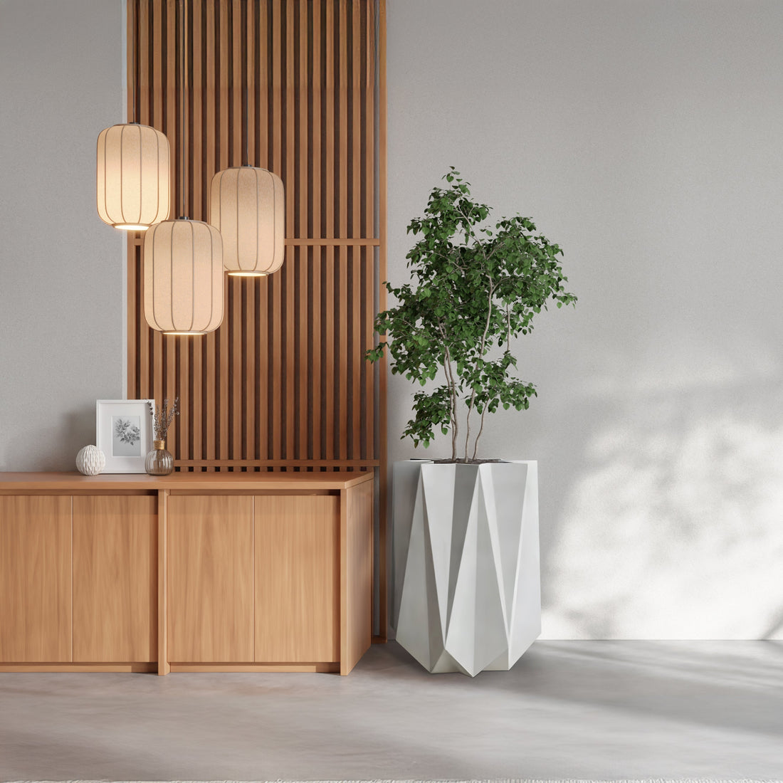 A minimalist interior features a wooden sideboard, slatted wall panels, and three pendant lamps highlighting a framed picture. Landmark Decors Visione Astratta Elegant Planter holds a leafy green plant, casting shadows on the light walls and floors to blend luxury with nature seamlessly.