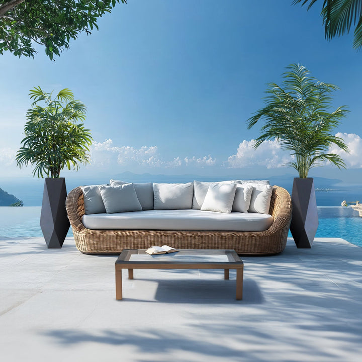 An outdoor patio showcases a wicker sofa with white cushions beside a pool. The scene is flanked by two Ombre Intrecciate Planters from Landmark Decor, merging luxury and nature. A glass-top coffee table sits in front, set against a backdrop of mountains and clear blue sky.