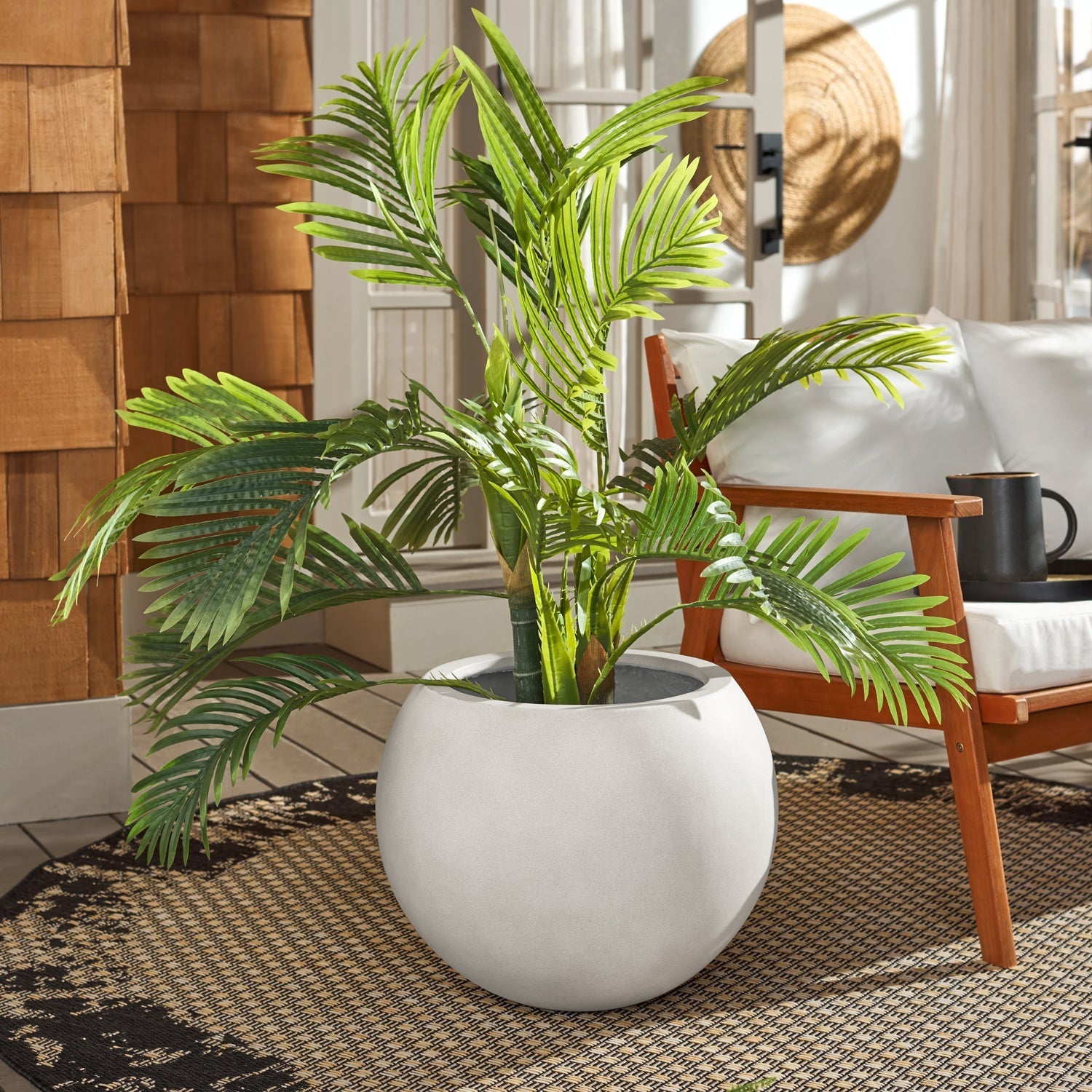 An Urban Vessel Planter by Landmark Decor, holding a large green palm, adorns a patterned rug beside a wooden armchair with white cushions. A cup sits on the armrest, and a sunhat hangs nearby, exuding luxury and nature in the bright, sunlit room.