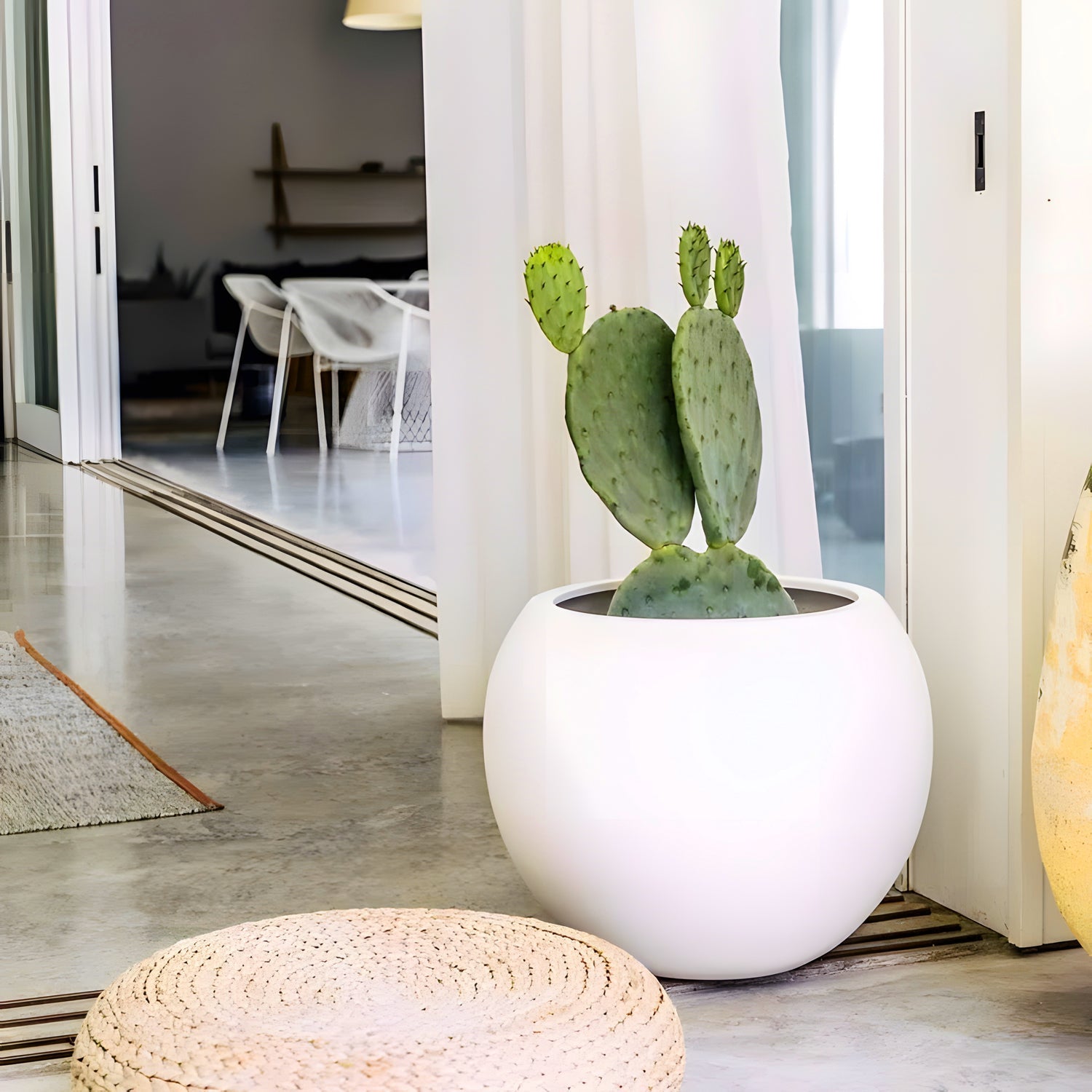 An Urban Vessel Planter by Landmark Decor, featuring a small cactus, is set on a concrete floor in a sleek, minimalistic interior. Nearby, a round woven cushion complements the scene, blending luxury with nature against the backdrop of a dining area with sleek white chairs.