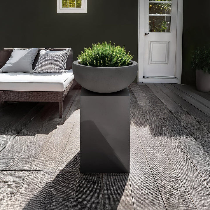A modern outdoor patio blends luxury and nature, featuring a dark wicker sofa with gray and white cushions. A sleek Obelisk Planter by Landmark Decor on a gray pedestal adds charm, complemented by wooden flooring and a white door in the background, achieving a minimalistic and elegant setting.