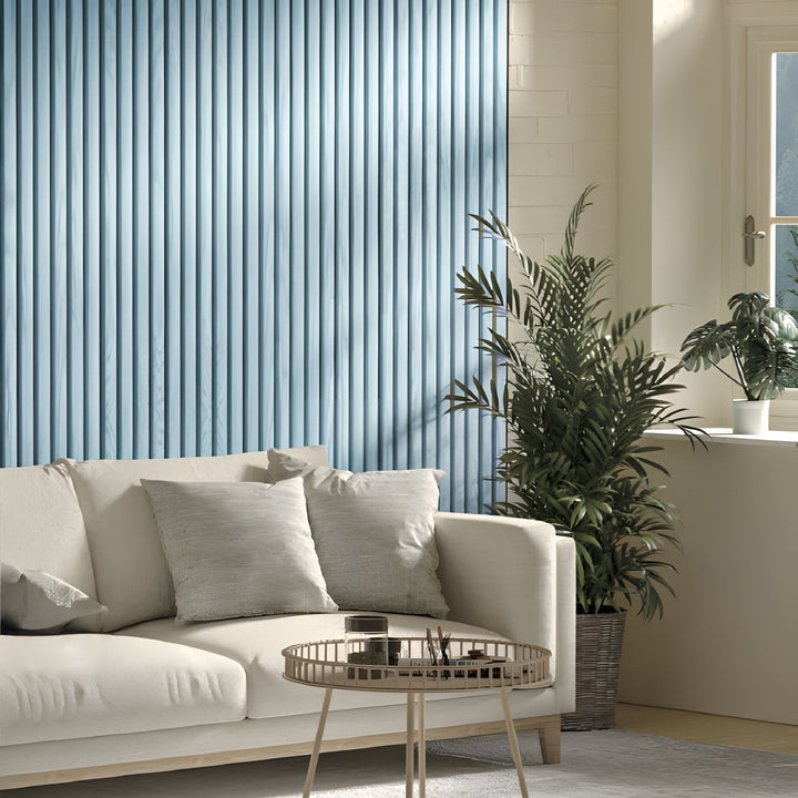 A beige sofa with two cushions is in a living room featuring Landmark Decors Sky Blue WPC Fluted Slat Wall Panel. A round coffee table sits in front, and a potted plant stands next to it. Natural light floods the room from a window with a plant on the sill.
