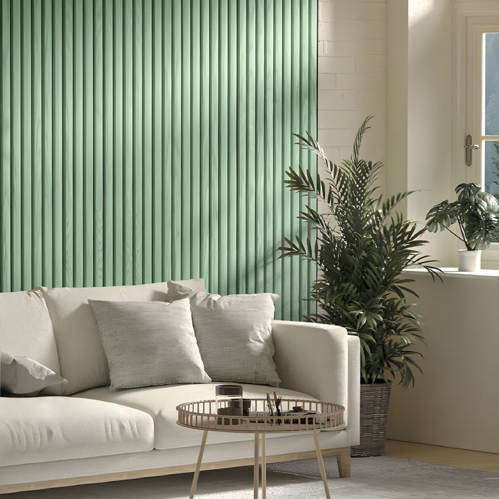 A modern living room showcases a light beige sofa with two gray cushions and a round coffee table. The calming ambiance is enhanced by a Mint Green WPC Fluted Slat Wall Panel from Landmark Decor, paired with a large potted plant and soft sunlight casting gentle shadows through the window.