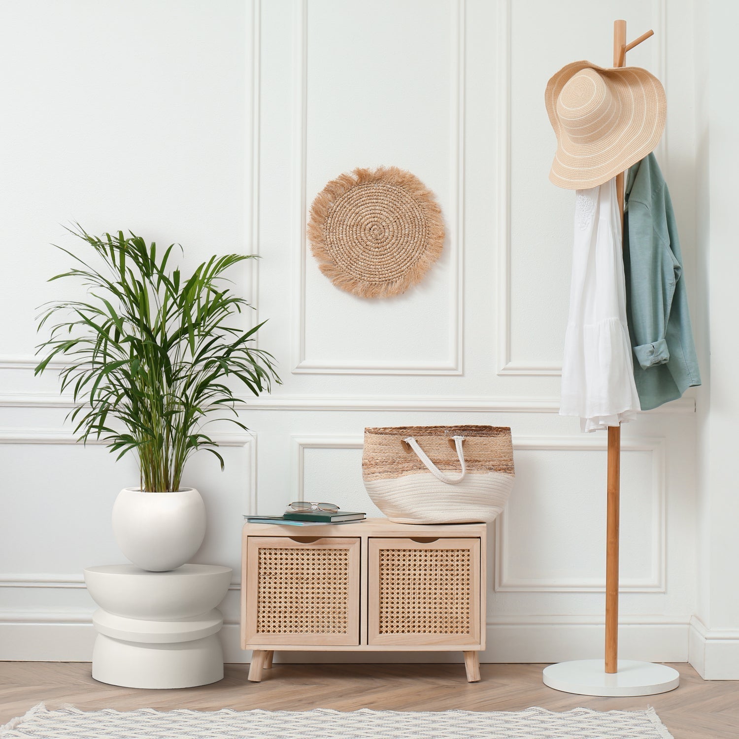 A stylish interior features the Landmark Decor Halo Harmony Stand and a wicker wall piece. A wooden coat rack holds a beige hat and clothing, while a small cabinet with woven drawers complements a handmade planter. Custom trees and a light-patterned rug enhance the rooms luxury and nature vibe.