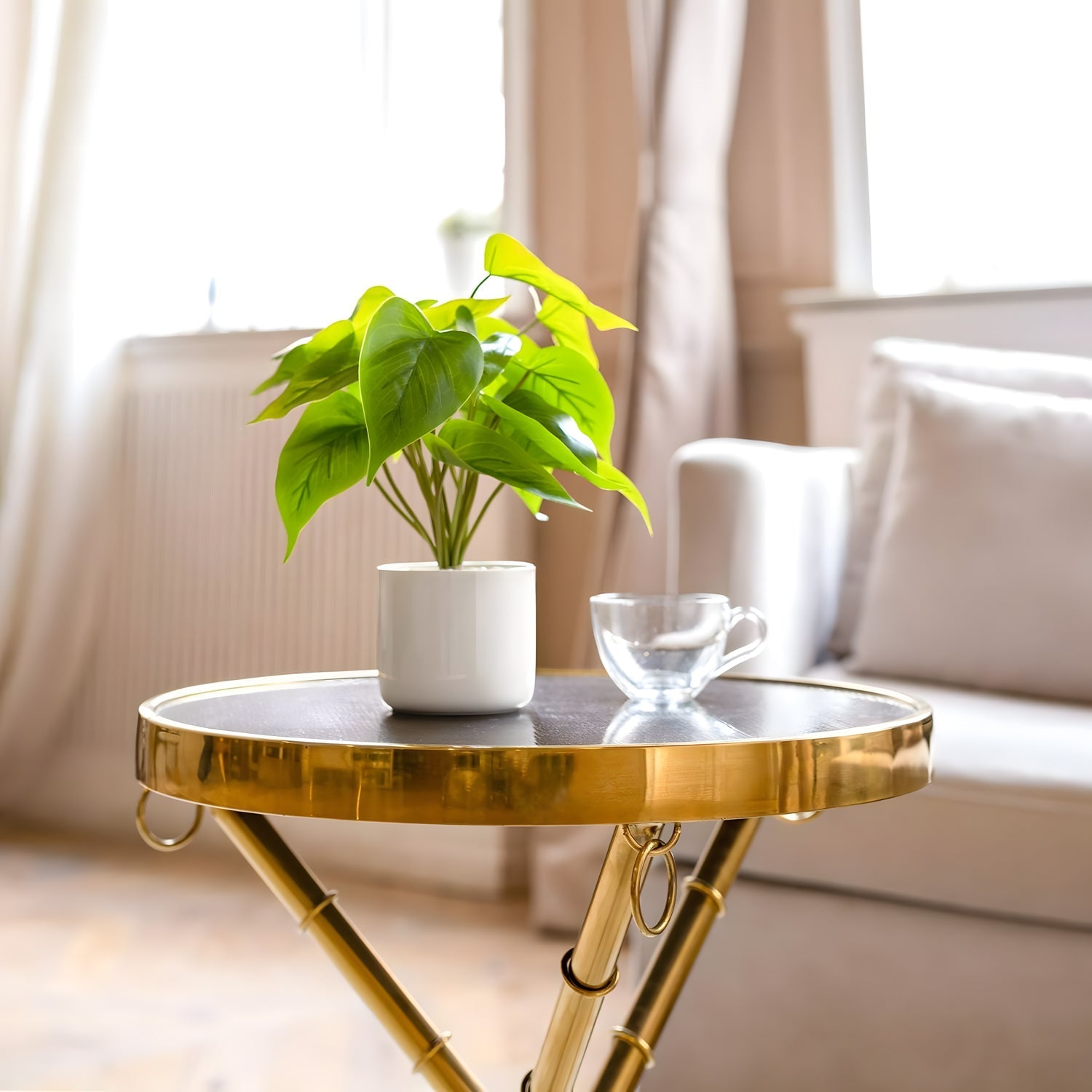 Landmark Decors Esmerald Heart, a round gold side table, holds a small pothos and a clear glass cup. It enhances a bright, cozy living room featuring a white sofa and large windows that flood the space with natural light.