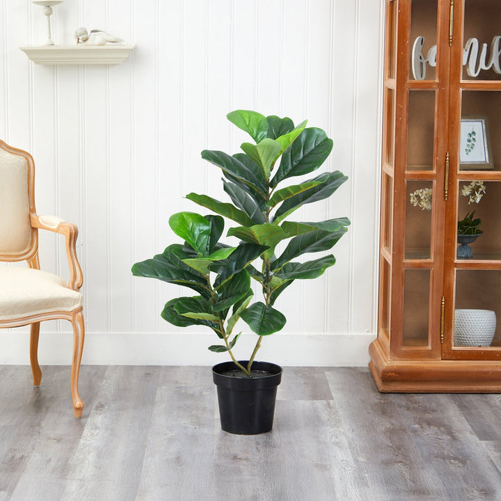 A medium Verdant Symphony plant by Landmark Decor graces the light wood floor, beside a cream chair and wooden cabinet with glass doors. A white paneled wall and decorative shelf backdrop enhance this inviting nook for the charming houseplant in its black pot.