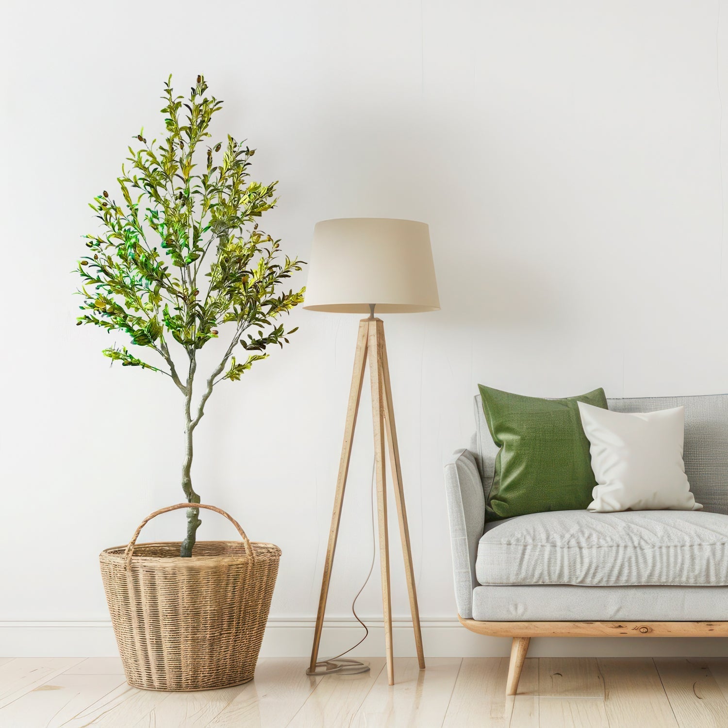 A cozy living room corner showcases Oliva Luxe, a long wicker basket with an elegant olive tree from Landmark Decor. It features a wooden tripod floor lamp with a beige shade and a light gray sofa adorned with green and white cushions, set against a white wall and light wood flooring.