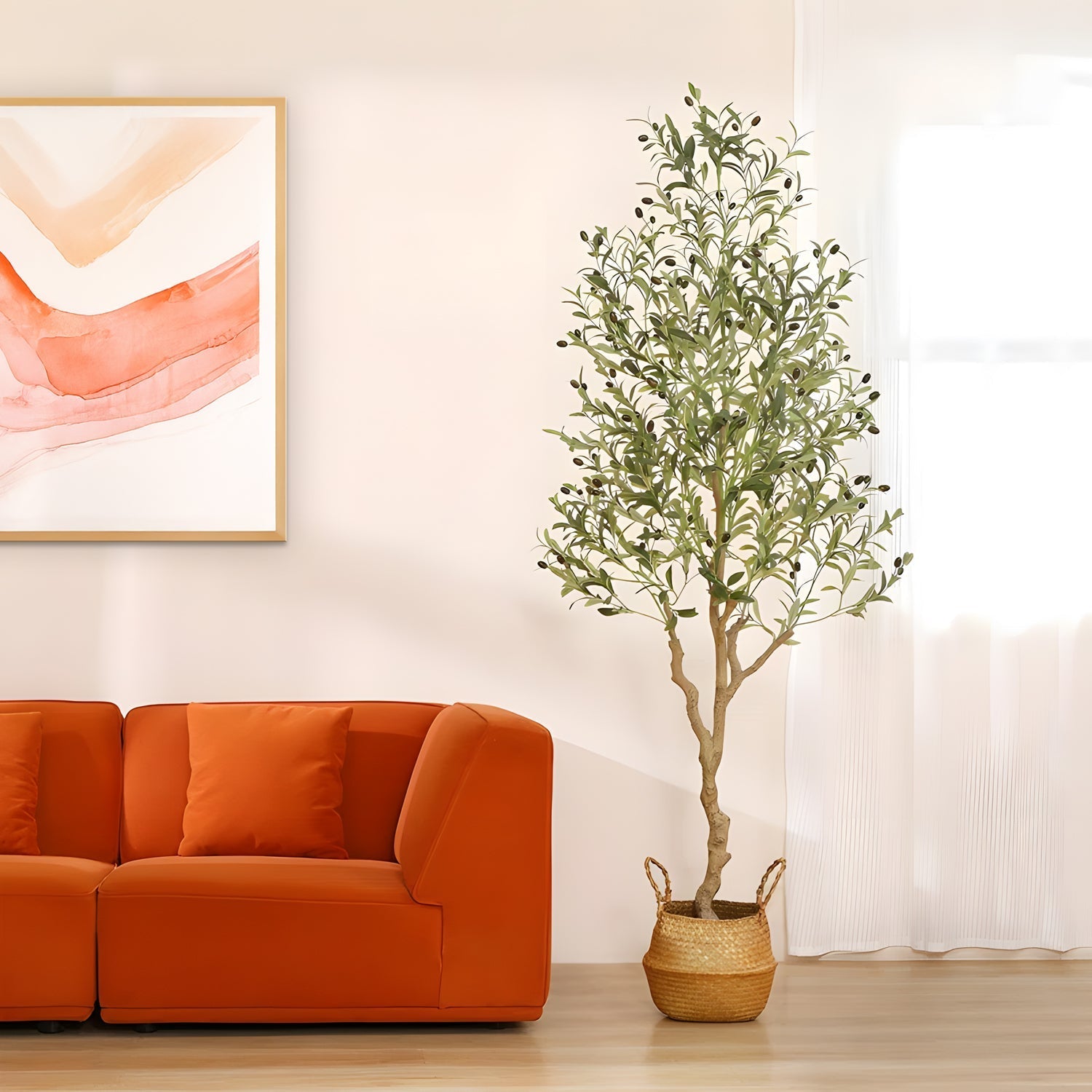 An elegant Oliva Luxe olive tree by Landmark Decor stands next to an orange sofa in a bright room. Above the sofa hangs a modern abstract painting with pink and orange hues. Natural light filters through a sheer white curtain on the right.