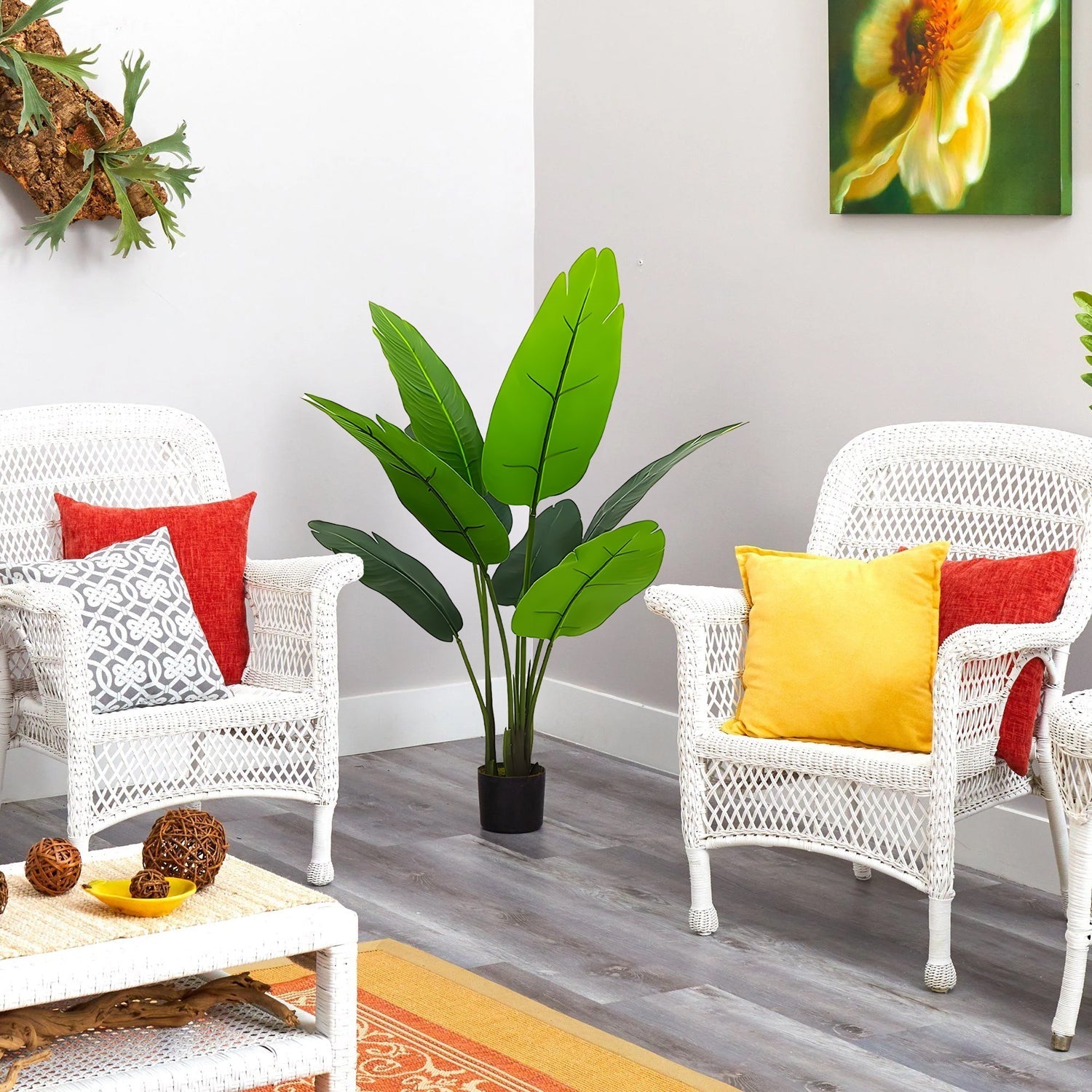 Two white wicker chairs with red and yellow cushions, a potted Banana Charm Strelitzia from Landmark Decor, and floral art on the walls create a serene elegance in a bright room with light wood floors. A table with decorative items is in the foreground.