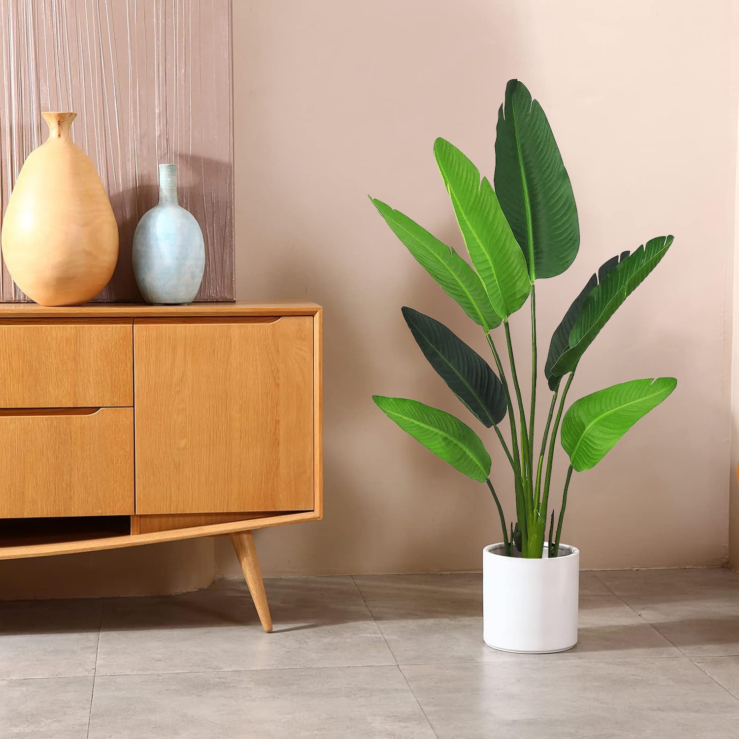A potted Banana Charm from Landmark Decor with lush green leaves is on a tiled floor near a wooden sideboard. The sideboard displays two vases, one beige and one light blue. A beige wall with vertical textured elements adds to the tranquil ambiance.