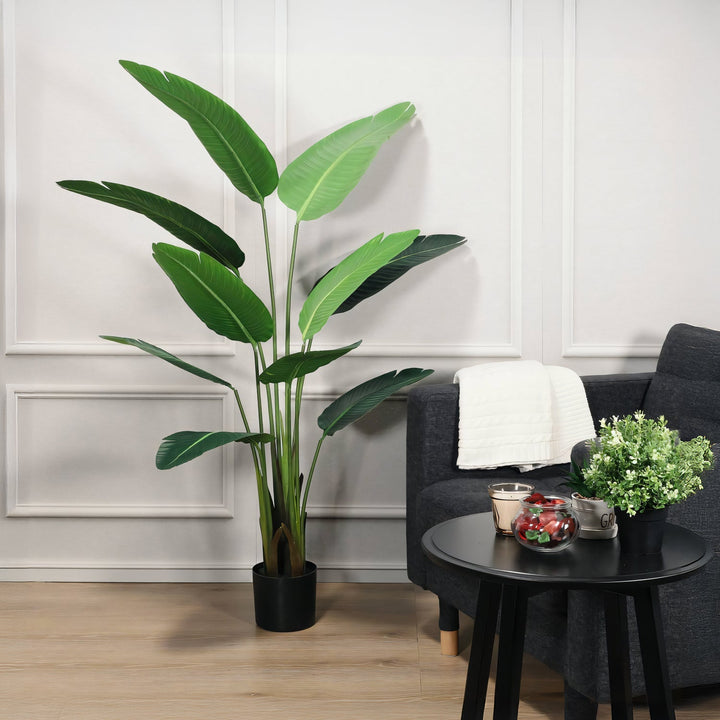 The Island Splendor, a tall green potted plant from Landmark Decor, sits beside a dark gray sofa and black round table. The table holds a small leafy plant, two mugs on a tray, and a glass jar with fruit against a white paneled wall backdrop.