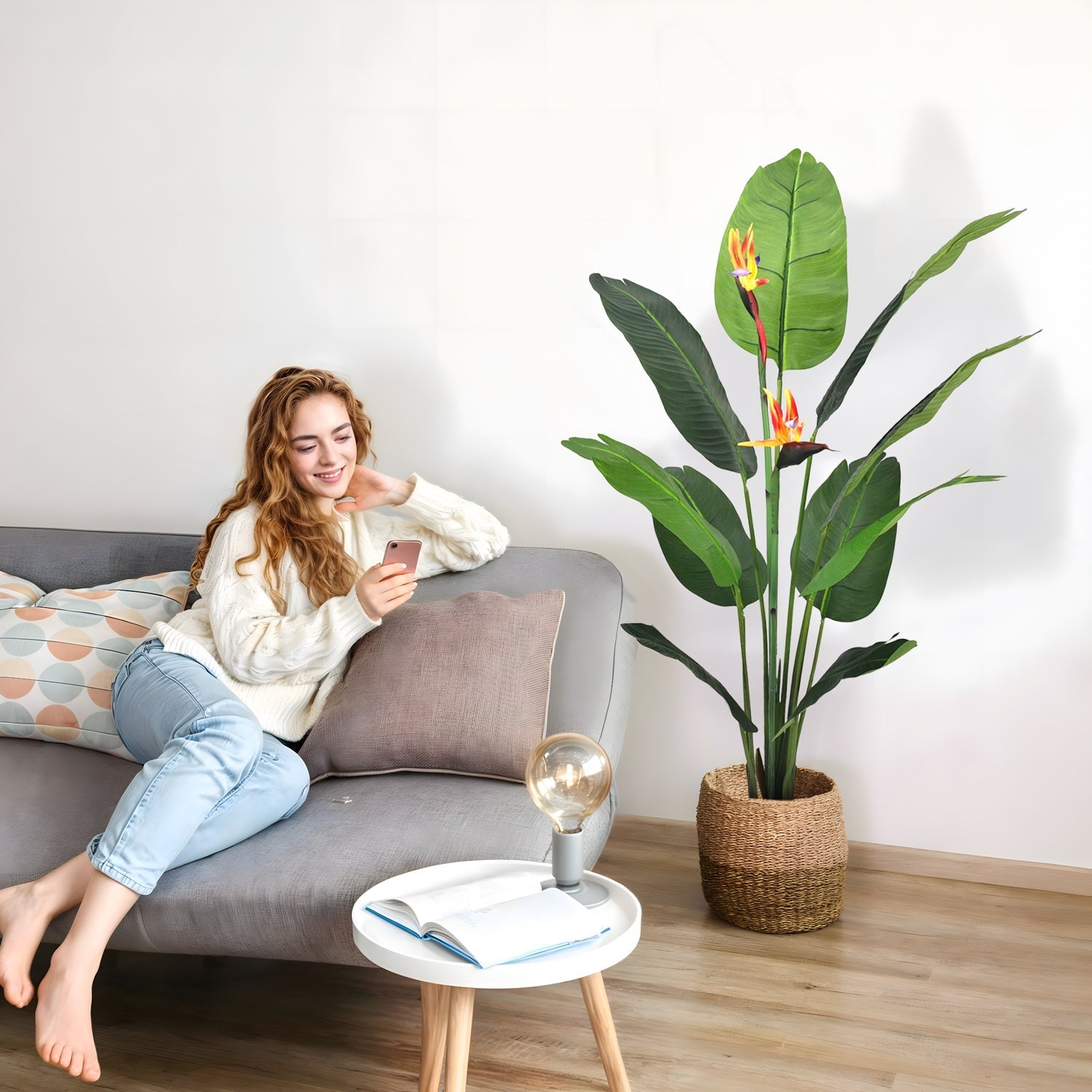A woman with wavy hair sits on a gray couch, looking at her phone, wearing a white sweater and light blue jeans. Beside her is the Bird of Paradise plant by Landmark Decor in a woven basket. A small white table with a book and lamp is in front.