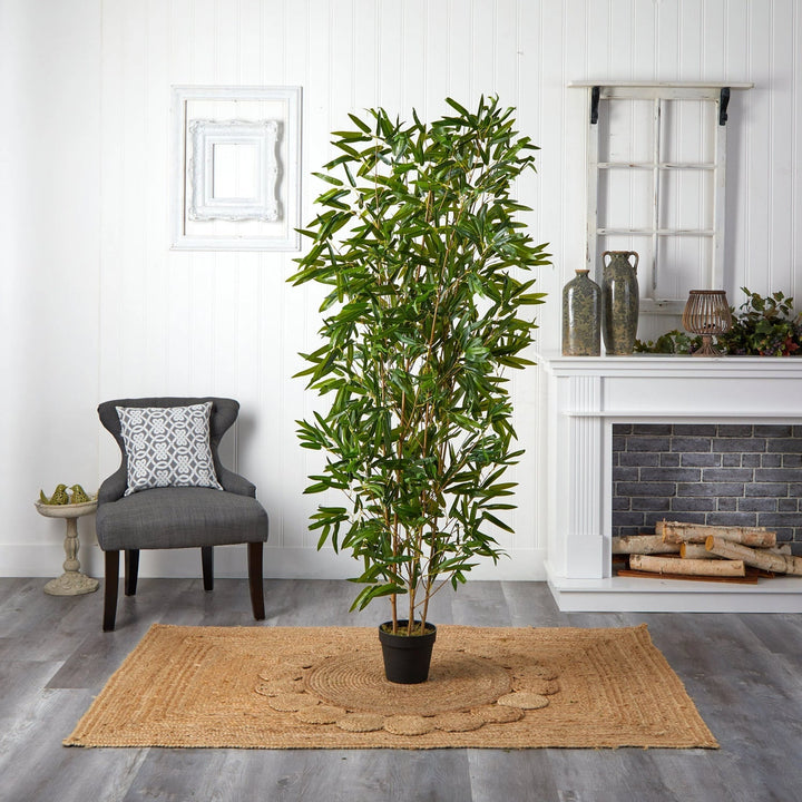 A cozy room features Landmark Decors Zen Whisper tall leafy plant on a woven rug. A gray armchair with a patterned cushion sits to the left, and the white mantelpiece to the right is adorned with decorative vases, while small logs nestle in the fireplace.