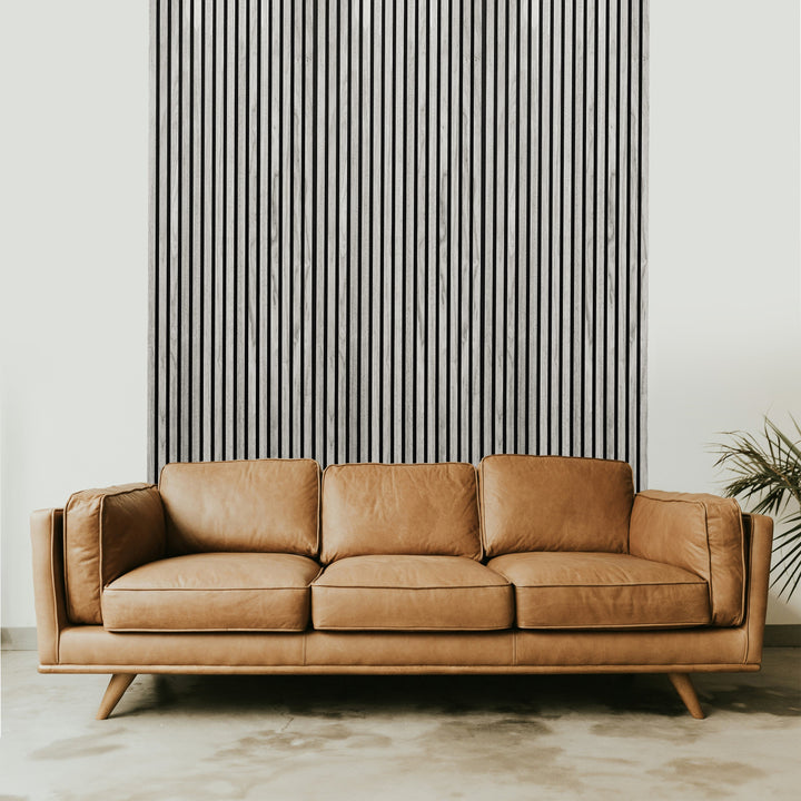 A brown leather sofa with wooden legs sits gracefully before a tall wall of Grey Oak Acoustic Slat Wall Panels from Landmark Decor, accentuating the modern and minimalist aesthetic. A small plant on the right enhances this luxury interior design settings natural charm.