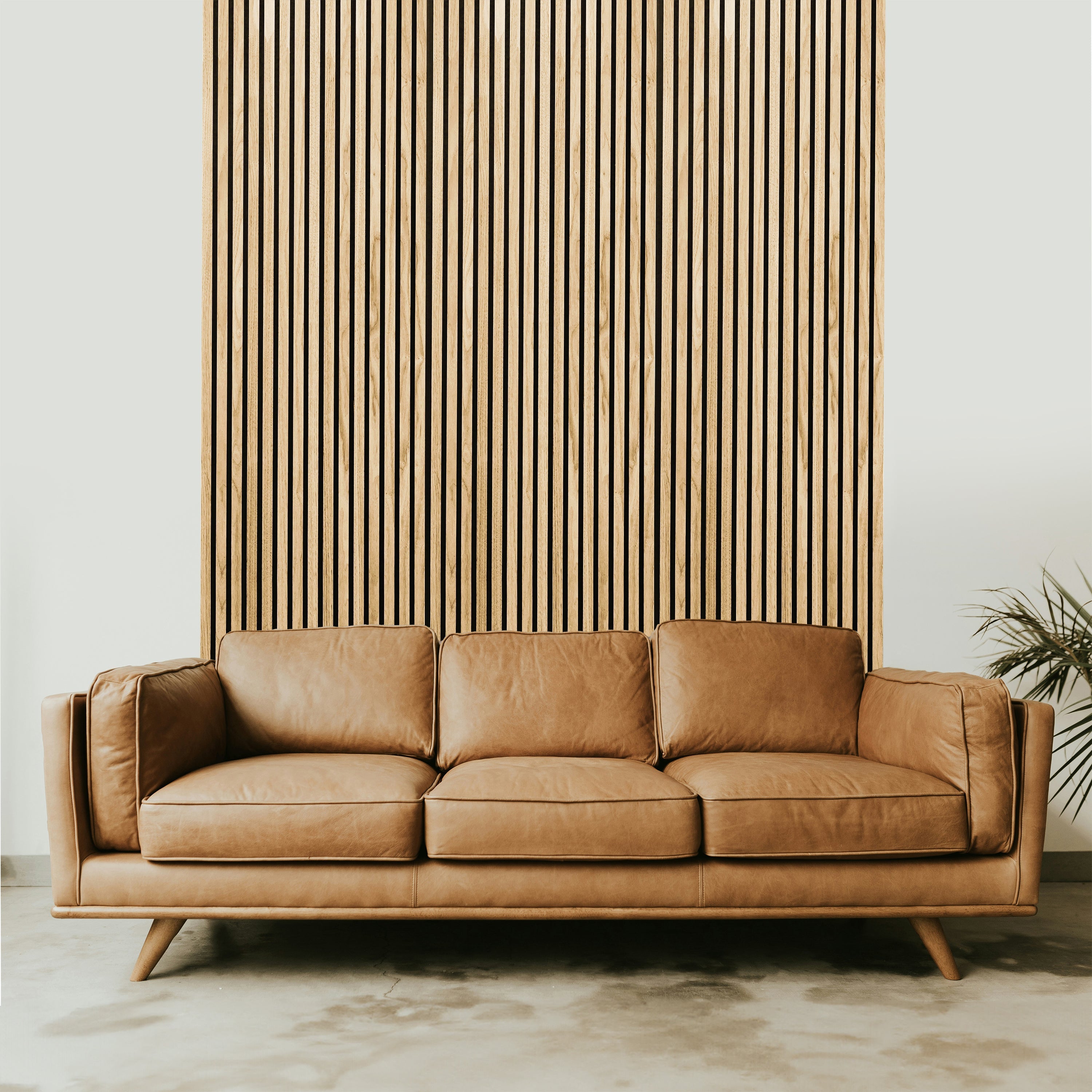 A tan leather sofa with three cushions sits against a Landmark Decor Natural Oak Wood Color Acoustic Slat Wall Panel, enhancing the luxurious interior. A green plant peeks from the right, complementing the neutral-toned polished floors sophistication.