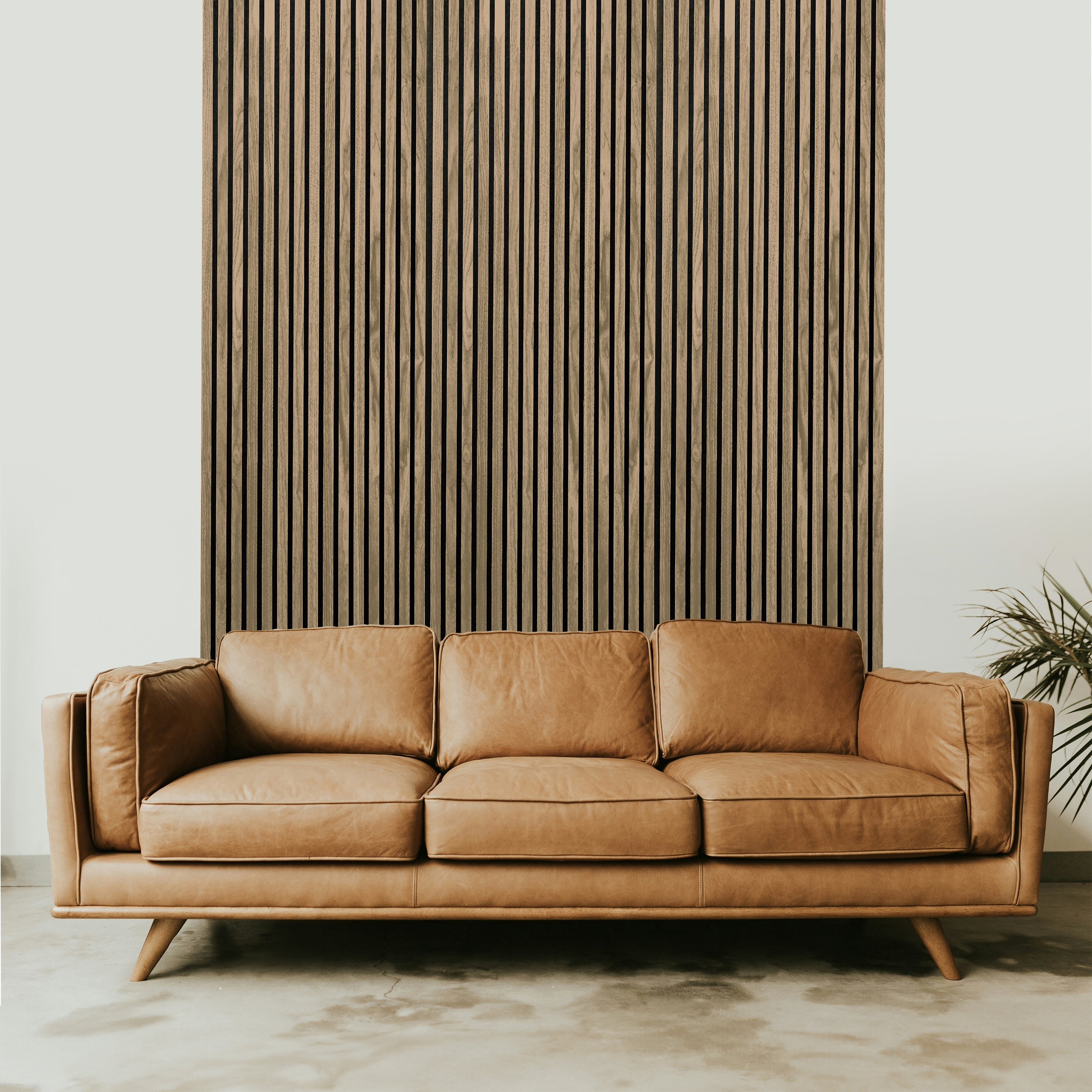 A brown leather sofa with three cushions is centered in front of a Landmark Decor Light Smoke Acoustic Slat Wall Panel, adding luxury to the design. A light-colored floor complements the setting while a small plant adds a touch of nature on the right side.