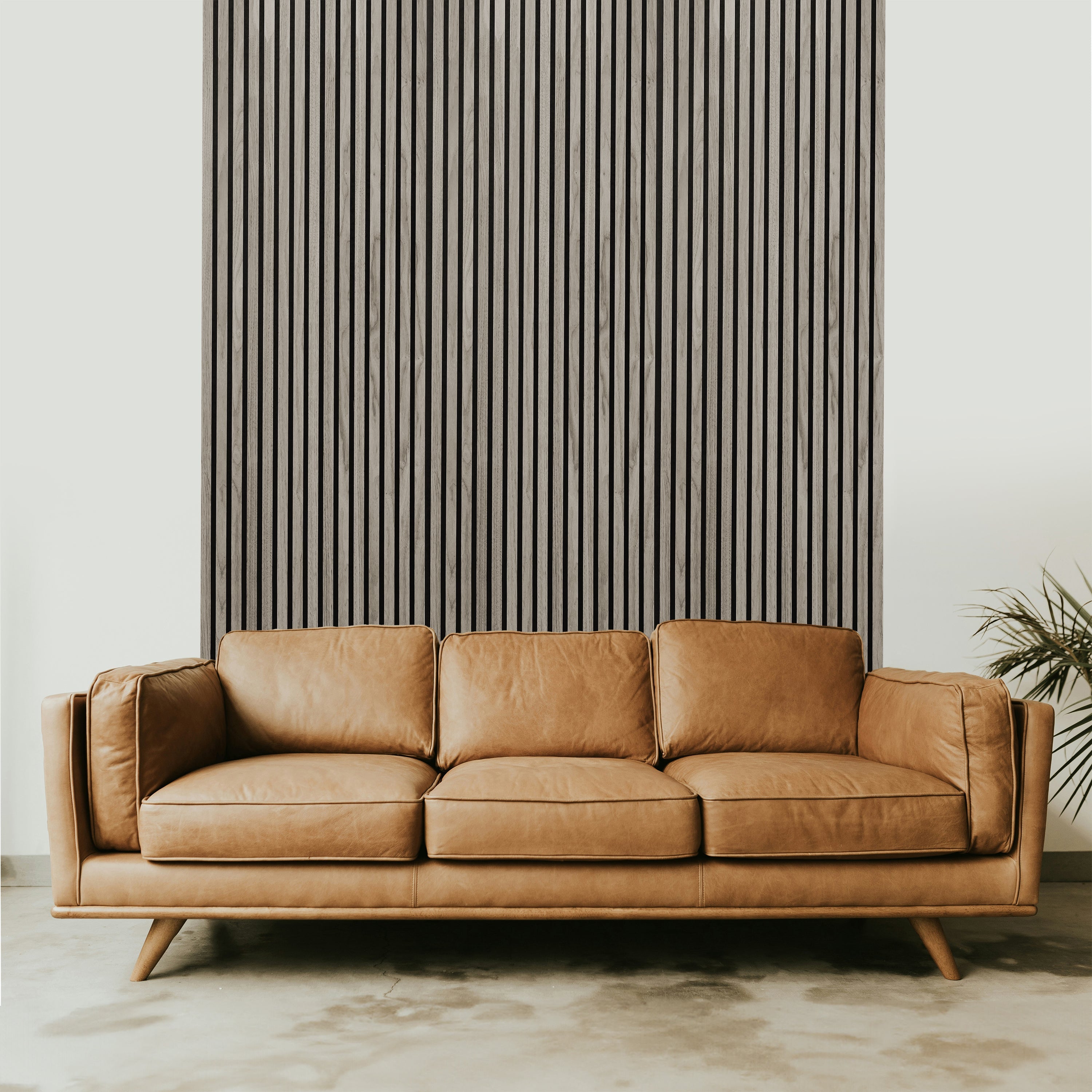 A brown leather couch takes center stage against a Landmark Decor Light Gray Acoustic Slat Wall Panel, exuding luxury. The rooms elegant design is complemented by a light-colored floor and a small plant, adding a touch of nature to the minimalist and modern ambiance.