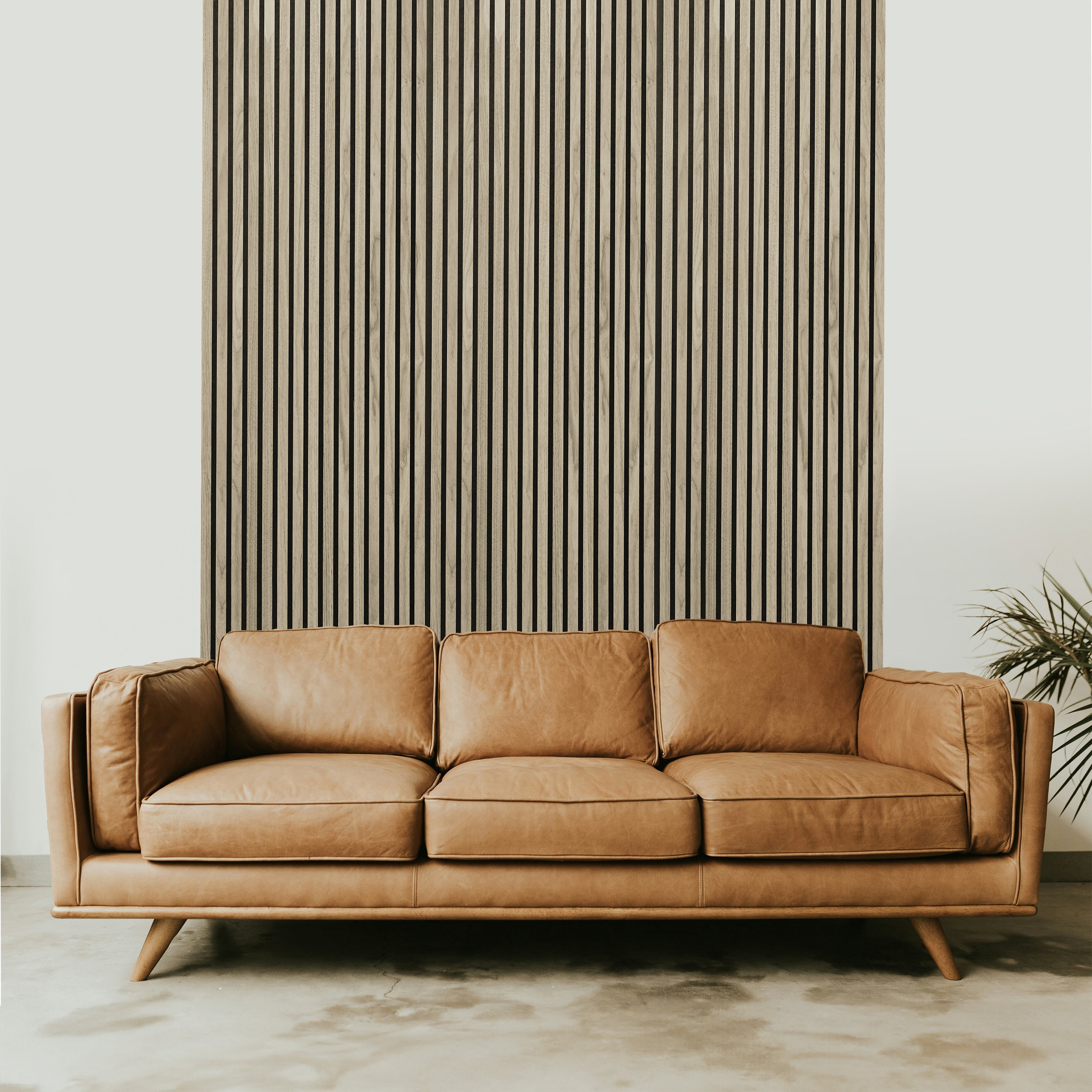 A tan leather sofa sits elegantly before a Landmark Decor Light Oak Acoustic Slat Wall Panel, adding warmth and texture. The room features smooth, light-colored concrete flooring, while a potted plant with slender leaves enhances the interior design from the right side.