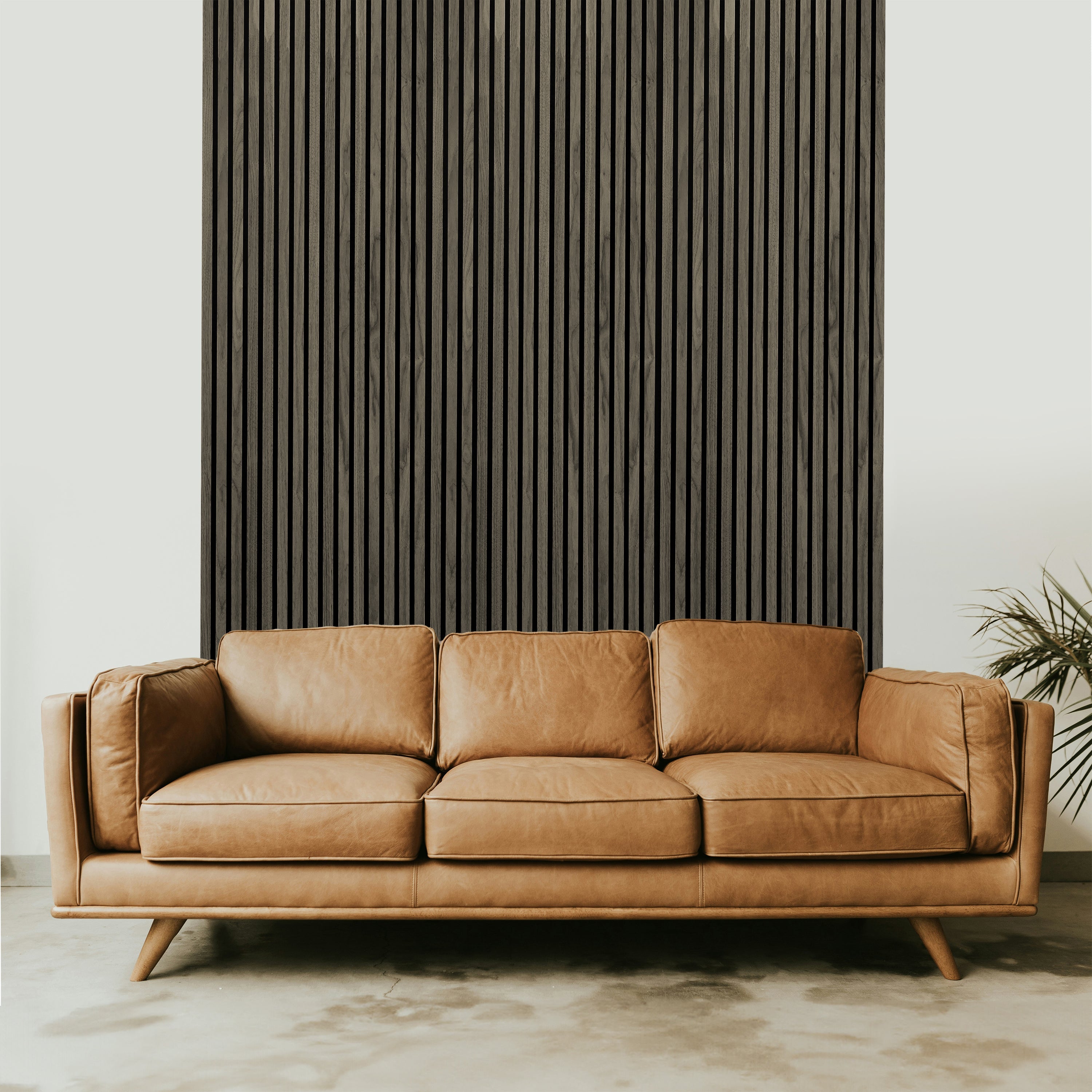 A brown leather sofa with three cushions sits on a light floor, backed by Landmark Decors Dark Gray Acoustic Slat Wall Panel adding sleekness. A small potted plant with thin green leaves to the right complements the sophisticated interior design.