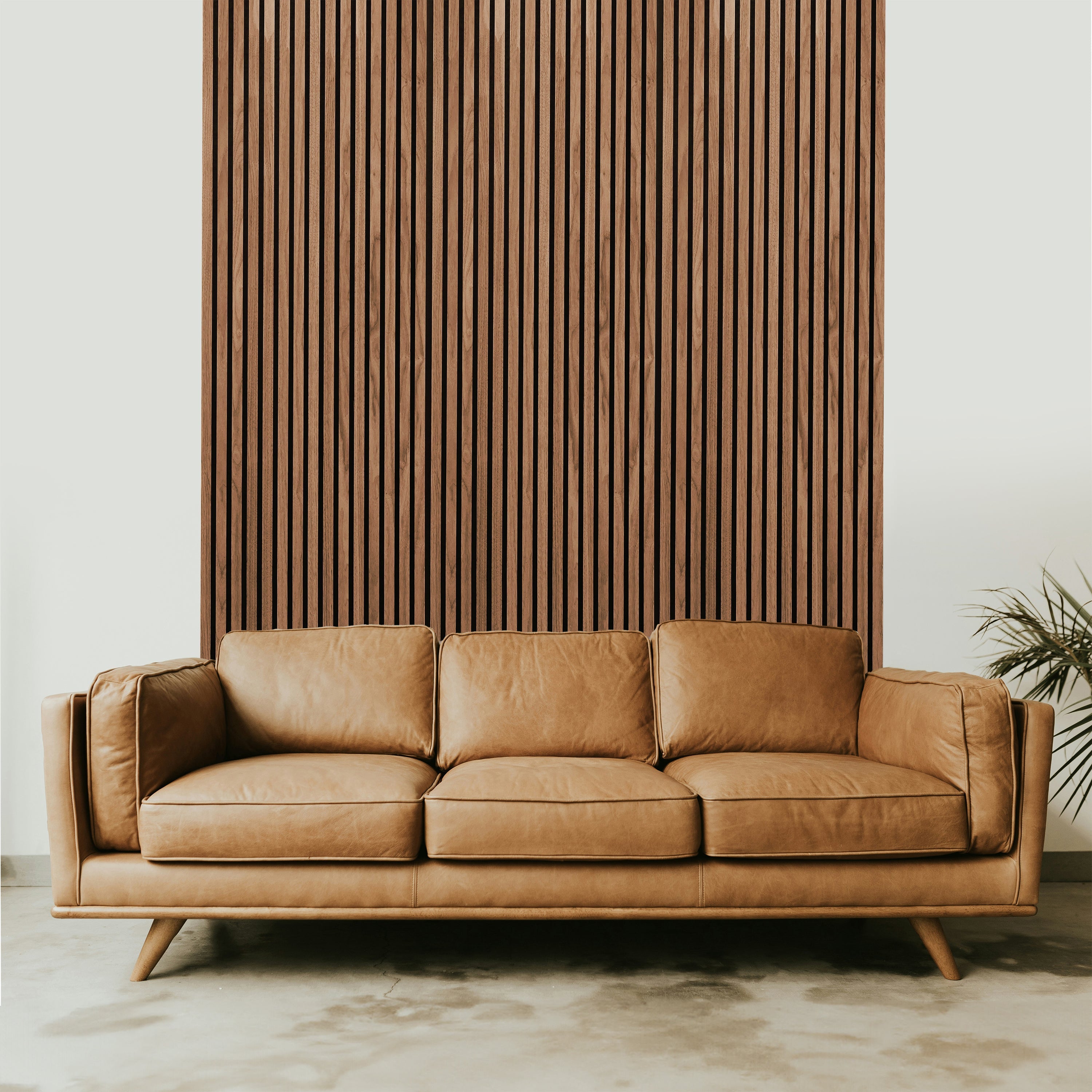A tan leather sofa rests against a wall adorned with Landmark Decors Light Smoke Oak Acoustic Slat Wall Panel. Featuring three seat cushions and angled wooden legs, it complements the elegant interior design, while a plant with long, narrow leaves adds a touch of nature on the right side.