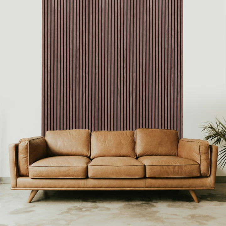 A brown leather sofa with wooden legs sits against a wall featuring Landmark Decors Dark Smoke Oak Acoustic Slat Wall Panel, enhancing the luxury interior. A green plant on the right adds a natural touch, while the room showcases a minimalist style with a light-colored floor.