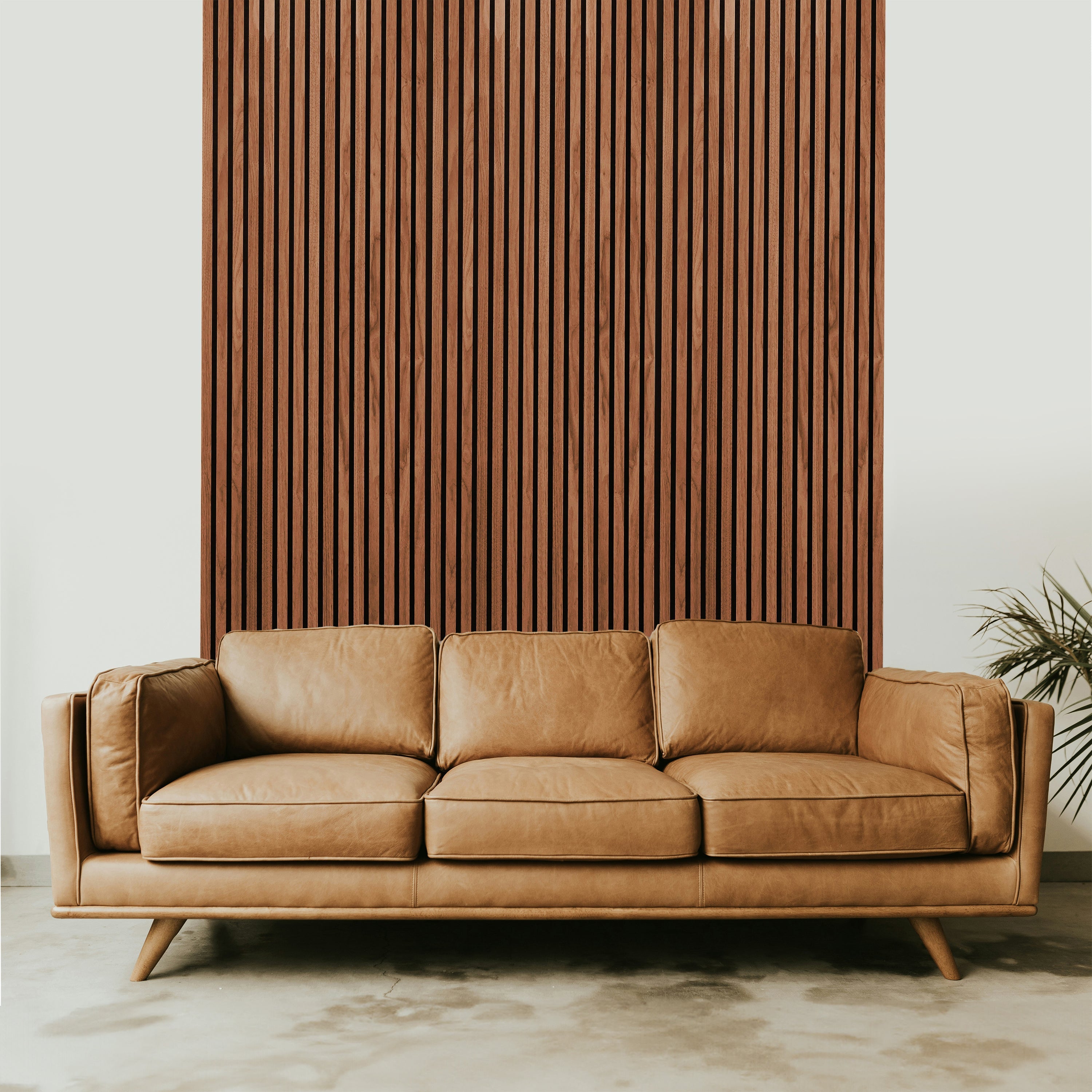 A minimalist, modern scene features a light brown leather sofa with three cushions on a concrete floor, framed by Landmark Decors Light Walnut Acoustic Slat Wall Panel. A plant with long, thin leaves peeks in from the right, creating a chic interior design.