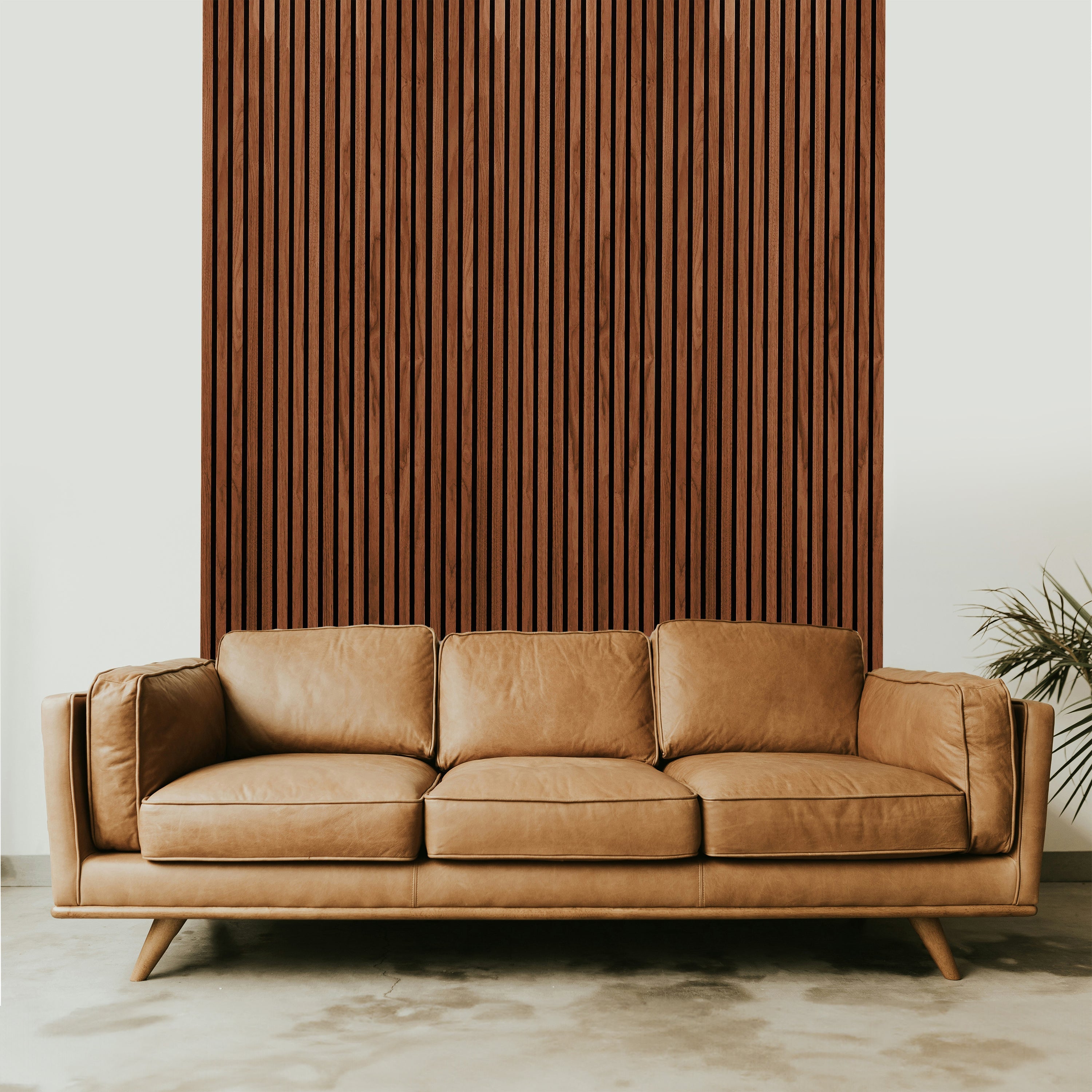 A tan leather sofa sits in front of a Landmark Decor Dark Walnut Acoustic Slat Wall Panel. A small plant on the right accents the minimalist design, creating a calm, modern interior flair.